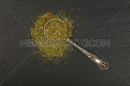 Close up one vintage antique metal spoon full of green dried herbs, dill or marjoram on background of black slate board, elevated top view, directly above