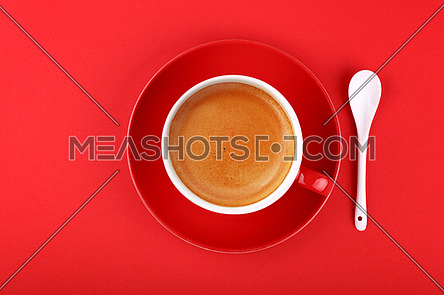 Close up one full cup of espresso coffee and saucer with white spoon over vivid red paper background, elevated top view, directly above