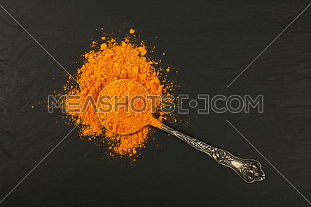Close up one vintage antique metal spoon full of yellow turmeric powder spice on background of black slate board, elevated top view, directly above