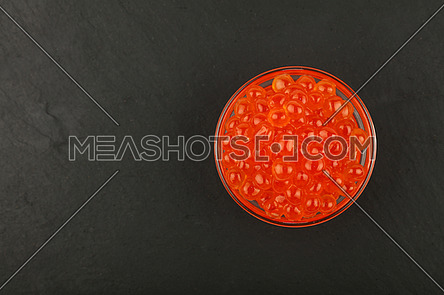 Close up one glass bowl of salmon fish red caviar on background of black slate kitchen board, elevated top view, directly above