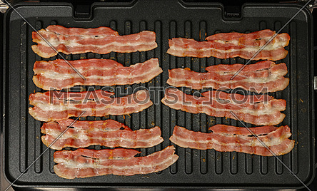 Close up cooking several bacon slices, rashers, on electric grill surface, elevated top view, directly above