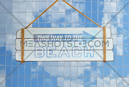 Close up old wooden directional arrow sign with this way to the beach text hanging over background of glass office building with reflection of blue cloudy sky
