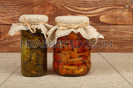 Close up of two glass jars of pickled green hot jalapeno chili peppers and brown honey fungus mushrooms with canvas top decoration and twine on tablecloth over brown wood background, low angle side view