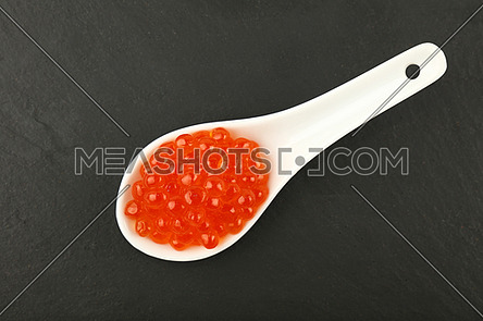 Close up one portion of salmon fish red caviar in white spoon on background of black slate kitchen board, elevated top view, directly above