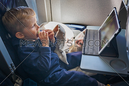 Baby boy is sitting on the plane and eats and looks at the tablet in front of him.