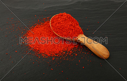 Close up one wooden scoop spoon full of red chili pepper or paprika powder on background of black slate board, high angle view