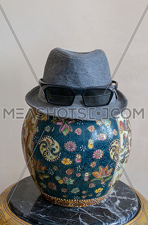 Gray trilby hat wearing sunglasses over antique decorated Chinese ceramic jar vase over antique black marble table