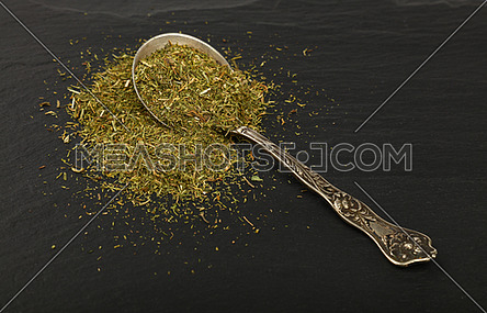 Close up one vintage antique metal spoon full of green dried herbs, dill or marjoram on background of black slate board, high angle view