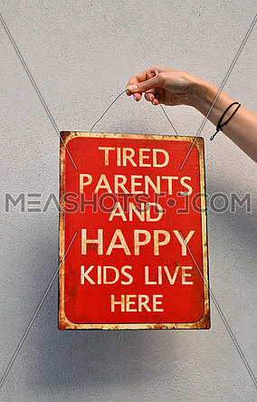 Woman hand holding red metal painted sign, tired parents and happy kids live here, over concrete wall with copy space