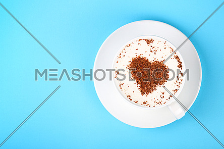 Close up one full white cup of frothy latte cappuccino coffee with heart shaped chocolate art over pastel blue paper background, elevated top view, directly above