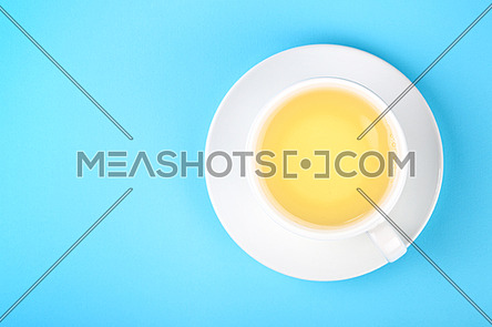 Close up one full white cup of green oolong or herbal tea on saucer over pastel blue paper background, elevated top view, directly above