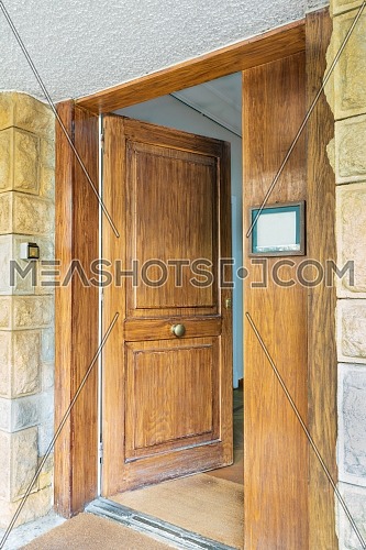 Half opened front wooden door at a stone bricks colored wall and a brown doormat