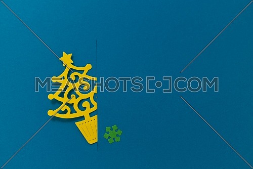 Small paper cut Christmas tree inserted into the slot on blue background with one green snowflake. Holiday postcard concept with copy space