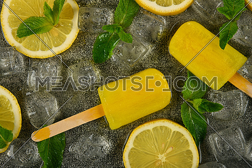 Close up frozen juice and limoncello popsicles with fresh lemon slices, green mint leaves and ice cubes on gray table surface, elevated top view, directly above
