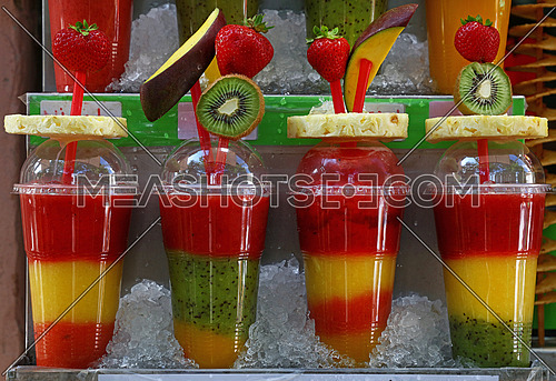 Close up selection of assorted fresh fruits, juices and mixed smoothies plastic glasses on ice at retail shelf, low angle view