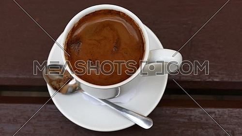 Close up one white cup full of Turkish black coffee and saucer on table with slow motion animated cinemagraph spin of coffee froth, elevated top view,