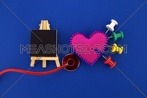 Medical concept with thumb tacks stuck into a felt heart symbolizing disease and illness with a red stethoscope below and small chalk board with copy space over blue