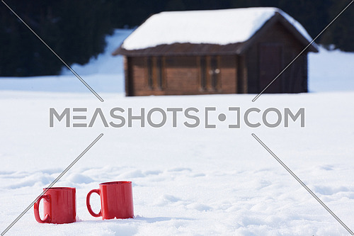 two red coups of hot tea drink in snow at beautiful winter  sunny day scene with wooden house in background in forest
