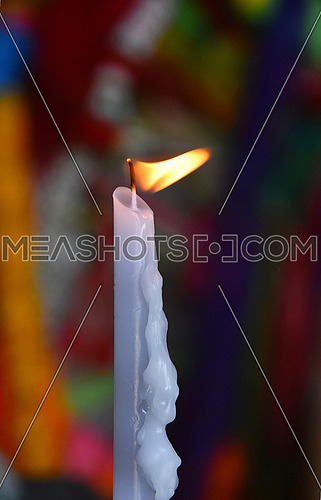 Burning white wax religious melting candle flame trembling in temple or church over vivid multicolor background