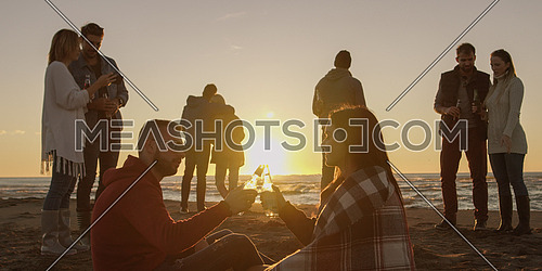 Happy Carefree Young Friends Having Fun And Drinking Beer By Bonefire On The Beach As The Sun Begins To Set