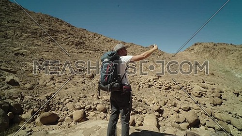 Follow shot for a male tourist wearing a pink cap and travel backpack Taking photos using mobile phone for Sinai Mountain for wadi Freij at day.