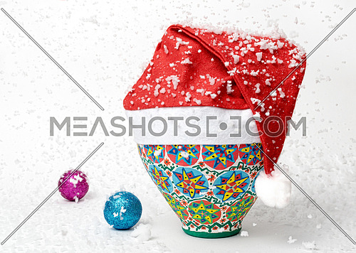 Egyptian decorated colorful pottery vase wearing Santa Claus red hat on white background with two colored shining Christmas balls and falling snow