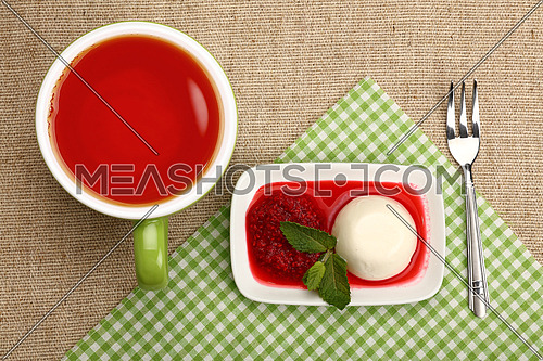 Close up serving panna cotta traditional Italian dessert with raspberry jam and cup of red black tea, elevated top view, directly above