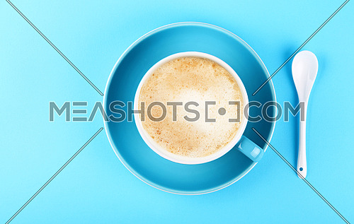 Close up one full cup of latte cappuccino frothy coffee and saucer with white spoon over pastel blue paper background, elevated top view, directly above