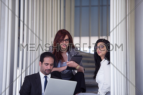 Young executives sitting on the steps of a corporate office building discussing business using laptop and tablet