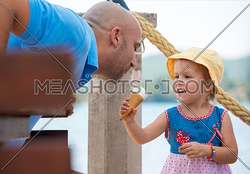 cute little girl having fun with her young father while eating ice cream by the sea during summer vacation holidays, celebration, children and people concept