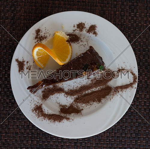 In the picture a slice of chocolate cake garnished with candied fruit, served on a white plate with a slice of orange and cocoa,view on above.
