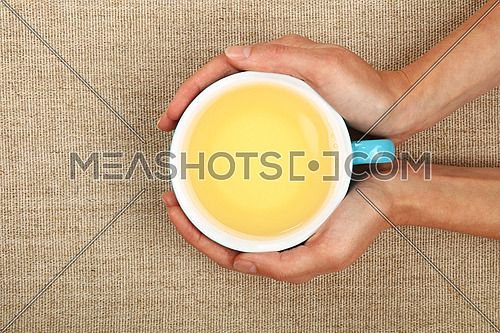 Two woman hands holding embracing big blue cup of clear green tea over linen canvas background, warming hands