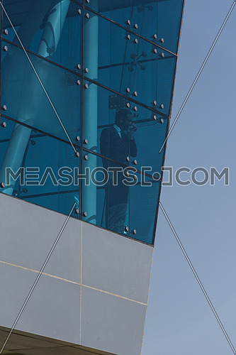 Outdoor shoot showing a young business executive talking over the phone through glass front of a corporate building with blue sky in background