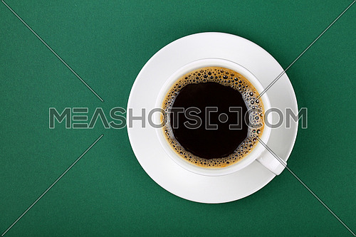 Close up one full white cup of black instant coffee on saucer on green table background, elevated top view, directly above