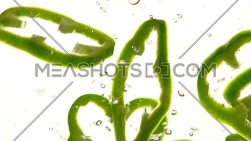 Close up several cut slices of fresh green bell pepper thrown and floating in clear transparent water, low angle side view, slow motion
