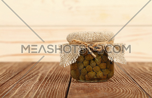 Close up of one small glass jar of pickled capers with canvas top decoration and twine on brown wooden table over white background, low angle side view