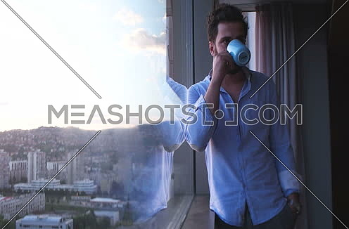 Man in shirt drinking coffee or tea by window admiring greath view on town during sunset