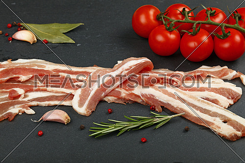 Raw pork bacon slices, rashers, spices, peppercorns, garlic, bay laurel leaves and red fresh cherry tomatoes on black slate board, close up, high angle view
