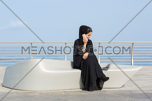 Saudi Lady wearing Black abaya making a call by the sea at day.