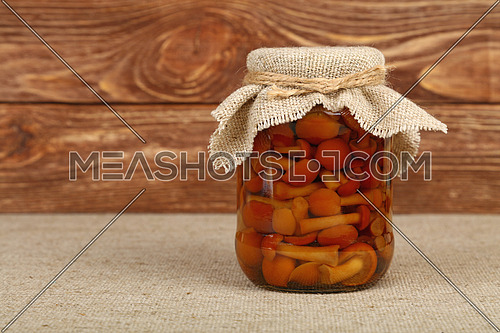Close up of one glass jar of pickled small brown honey fungus Armillaria mushrooms with canvas top decoration and twine on canvas tablecloth over brown wooden background, low angle side view