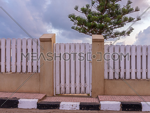 White weathered wooden garden gate and fence with background of single tree and cloudy sky at Montaza public park, Alexandria, Egypt