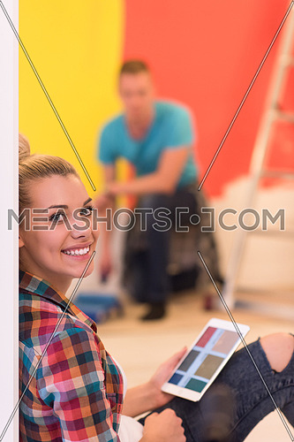 Happy couple doing home renovations, the man is painting the room and the woman is relaxing on the floor and connecting with a tablet