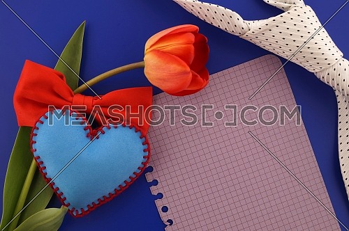 Blank notepaper for a Valentines message with red tulip, handmade blue felt heart, a bowtie and necktie over a colorful blue background