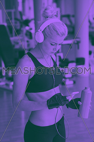 young healthy woman drinking water  in fitness gym while sitting on pilates ball and listening music on headphones from smartphone duo tone