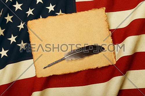 Close up blank vintage yellow paper sheet and quill on old weathered cotton embroidered US national flag background, symbol of American history, elevated high angle view, directly above