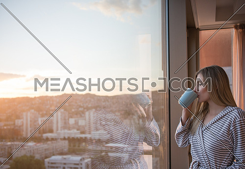 young woman in bathrobe enjoying evening coffee and beautiful sunset landscape of the city while standing by the window
