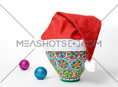 Studio shot of Egyptian decorated colorful pottery vase wearing Santa Claus red hat on white background with two colored shining Christmas balls