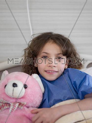 young girl lies in a hospital bed while hugs  pink teddy bear portrait