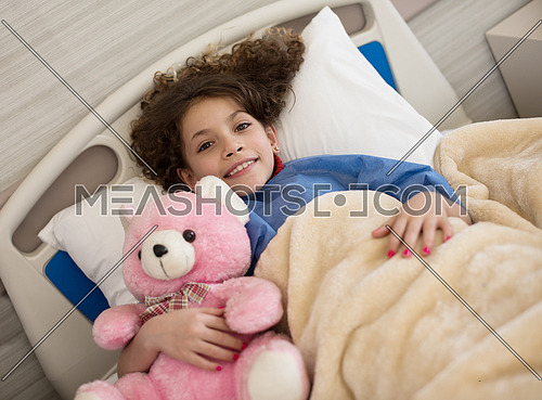young middle eastern girl lies in a hospital bed while hugs pink teddy bear
