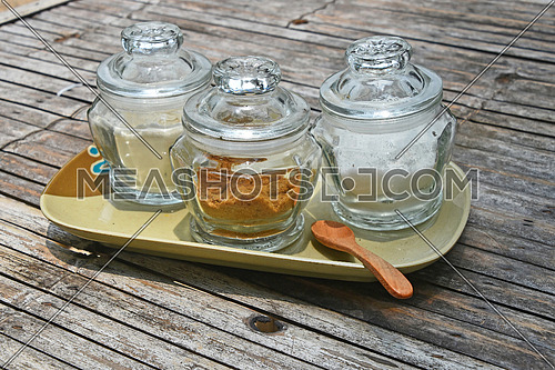 Three glass jars of different sugar, white and brown cane, on ceramic saucer with wooden spoon on old vintage bamboo table for coffee time